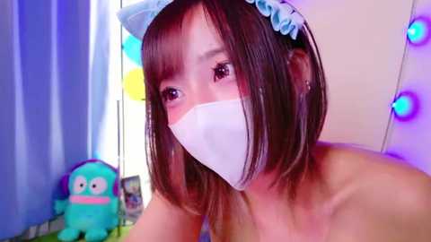 Media: A video of a young Asian woman with shoulder-length brown hair, wearing a light blue cat headband, a white surgical mask, and no top, set against a colorful, festive background with fairy lights and a plush toy.