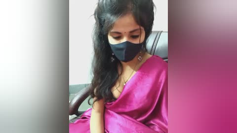 Media: Video of a South Asian woman with long, dark hair, wearing a magenta sari and a black face mask, seated indoors, with a blurred background.