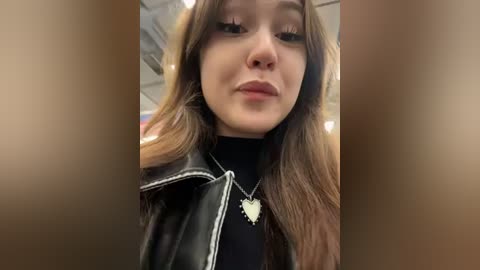 Media: Video of a young woman with light skin and long, wavy brown hair, wearing a black leather jacket over a black turtleneck, and a silver heart necklace. She has a neutral expression.