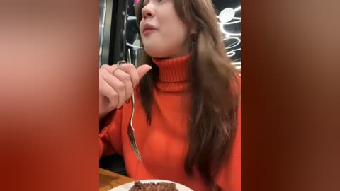 Media: Video of a young woman with long brown hair, wearing a bright orange turtleneck sweater, holding a fork, seated at a wooden table in a dimly lit restaurant.