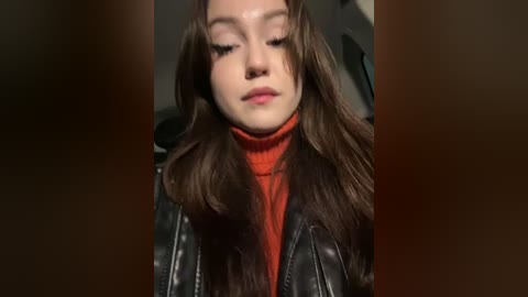 Media: Video of a young woman with long brown hair, wearing a red turtleneck sweater under a black leather jacket, with a blurred background.