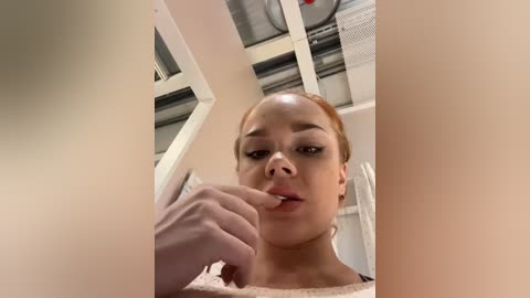 Media: Video of a young woman with light skin and red hair, wearing a beige sweater, touching her lips, taken from a low angle in a laundry room with exposed pipes and a metal vent.