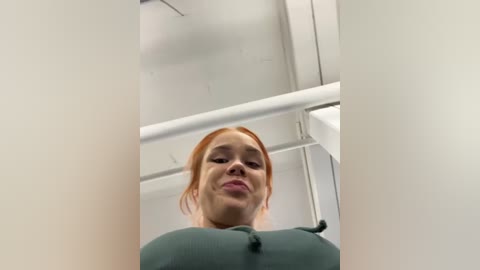 Media: Video of a young woman with light skin and red hair, wearing a green top, making a pouty face. Background features a white ceiling and a metal railing.