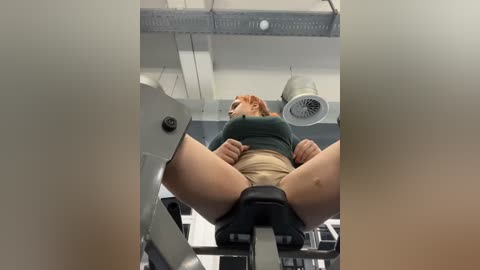 Media: A video of a young, fair-skinned woman with red hair, wearing a green top and beige shorts, lying on a gym bench with her legs spread, in a gym with metallic equipment.