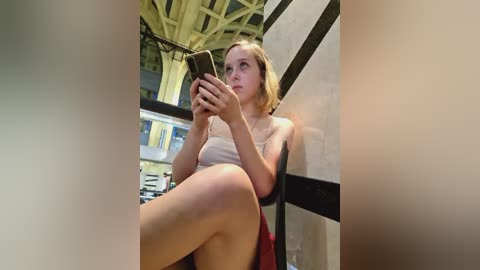 Media: A video of a fair-skinned woman with short blonde hair, wearing a white tank top and black skirt, sitting on a chair, engrossed in a smartphone, in a modern, glass-walled room with a high, arched ceiling.