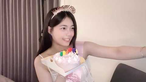 Media: Video of a young Asian woman with long black hair, wearing a pink princess tiara and a white, sleeveless dress, smiling and holding a birthday cake with colorful candles. Background includes gray curtains and a beige wall.