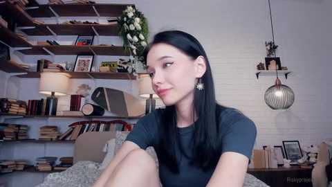 Media: Video of a young woman with straight black hair, wearing a black t-shirt, sitting on a bed in a cozy, book-filled room with white brick walls and hanging lamps.