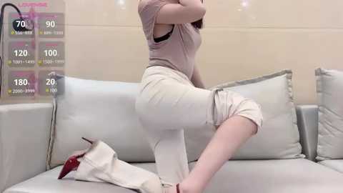 Media: Video of a light-skinned woman in a beige top and white pants, squatting on a white sofa, with a white handbag and high heels nearby.