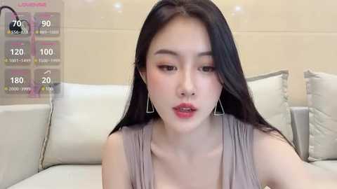 Media: A video of an Asian woman with long black hair, wearing a sleeveless gray top, sitting on a white couch. The background shows a beige wall and a digital temperature display.