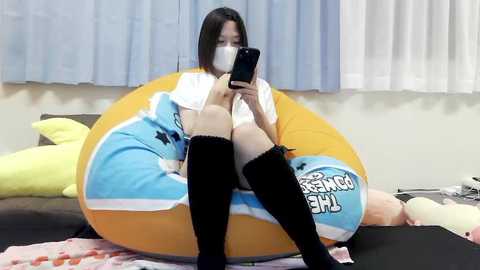 Media: Video of an Asian woman with long black hair, wearing a white mask, black knee-high socks, and a blue and yellow plush lounge chair. Background includes a bed, pillows, and white curtains.