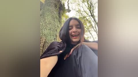 Media: A video of a young woman with light brown skin, long dark hair, and a black cloak, smiling widely outdoors in a forest, surrounded by trees.