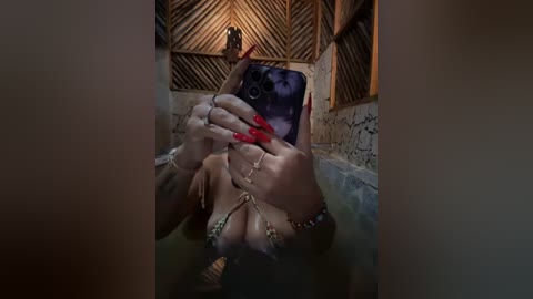 Media: Video of a woman with fair skin, long red nails, and a green bikini top taking a selfie in a wooden-paneled, dimly lit bathroom with a blue-tiled tub.