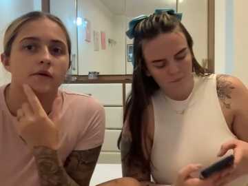 Media: Video of two women in a bathroom; one with light skin, pink shirt, and tattoos; the other with darker skin, long brown hair, and a white tank top, both looking at a smartphone.