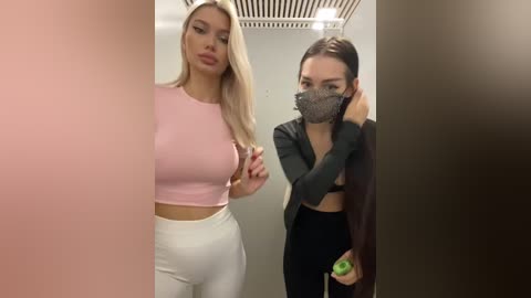 Media: Video of two women in a modern, well-lit room: one in a pink crop top and white leggings, the other in a black blazer and mask, adjusting her hair.