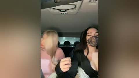 Media: Video of two women wearing black face masks in a car, blurred background. One has long blonde hair, the other has long dark hair.