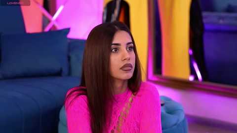 Media: Video of a young woman with long, straight brown hair, wearing a bright pink lace top, sitting on a teal sofa in a modern, brightly lit room with vibrant, neon-colored walls.