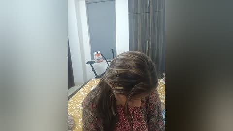 Media: Video of a young woman with long, wavy brown hair, wearing a patterned sweater, sitting on a bed with a yellow floral comforter in a dimly lit, sparsely furnished room.