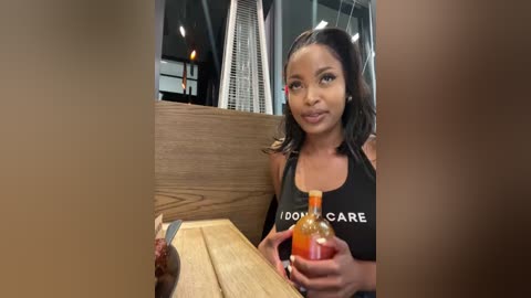 Media: Video of a young Black woman with medium brown skin, wearing a black tank top with white text, sitting at a wooden table in a dimly-lit restaurant. She holds a bottle of hot sauce, smiling.