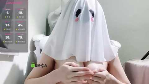 Media: Video of a topless woman with pale skin, wearing a white sheet with cartoon ghost eyes covering her head, holding her breasts. Background includes a white chair and a screen showing a fitness app.