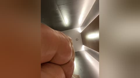 Media: A video showing a close-up view of a person's bare, large buttocks and a hand holding them, set in a modern, minimalist room with angled, light-emitting ceiling panels.