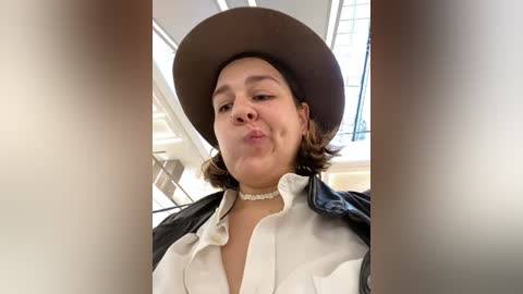 Media: Video of a young woman with fair skin and short brown hair, wearing a white shirt, black jacket, and a wide-brimmed brown hat, making a pouty face. She's indoors, with bright natural light streaming in.