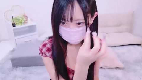 Media: Video of an Asian woman with long black hair, wearing a pink surgical mask, red tie-dye top, and white fuzzy socks, kneeling on a light grey carpet, in a minimalist bedroom with white walls and a bed.