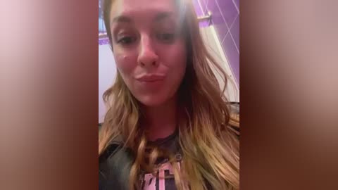 Media: Video of a Caucasian woman with long, wavy blonde hair, wearing a black graphic t-shirt, making a pouty face. Background shows a tiled bathroom with purple accents.