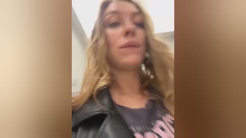 Media: Video of a young woman with long blonde hair, wearing a black leather jacket and a dark T-shirt with pink lettering. She has a neutral expression and stands indoors with a blurred background.