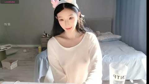 Media: Video of an Asian woman with long black hair, wearing a white long-sleeve shirt and pink cat headband, sitting in a minimalist bedroom with a bed, white sheets, and a blue curtain.