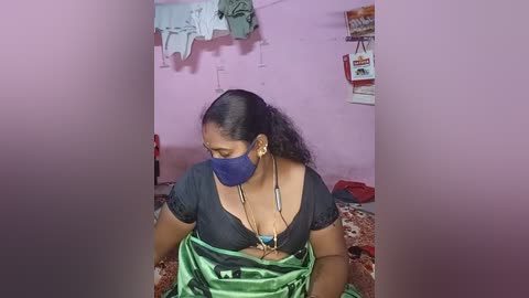 Media: Video of a South Asian woman with medium brown skin, wearing a black mask, green saree, and blue blouse, seated in a pink-walled room with laundry hanging and a bag on a wall.