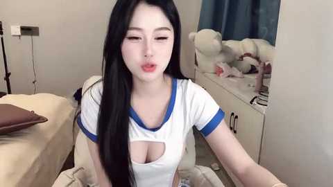 Media: Video of a young Asian woman with long black hair, wearing a revealing white sports top with blue trim, in a bedroom with a bed, stuffed animals, and a mirror.