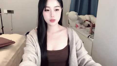 Media: A video of a young Asian woman with long black hair, wearing a brown top and a light gray cardigan, sitting on a bed in a bedroom with plush toys and a mirror.