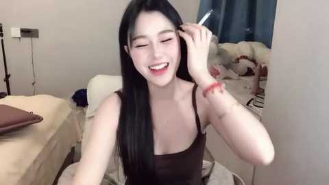 Media: Video of a smiling Asian woman with long black hair, wearing a black spaghetti strap top, applying makeup in a bedroom with a bed and stuffed animals.