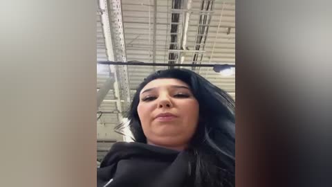 Media: A video of a young woman with long black hair and a serious expression, taken from a low angle inside a warehouse with a metallic ceiling and exposed pipes.