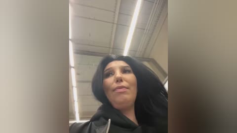 Media: Video of a woman with long black hair, wearing a black hoodie, standing in an office with fluorescent lighting. She has a neutral expression and is looking upwards.