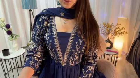 Media: Video of a woman in a blue embroidered dress with a scarf, sitting on a chair in a cozy room with white curtains, a lamp, and decorative plants.