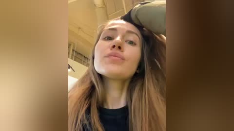 Media: Video of a Caucasian woman with straight, light brown hair, wearing a black top, blowing a kiss. Background shows an indoor, beige, industrial ceiling with exposed pipes.