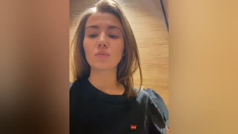 Media: Video of a young Caucasian woman with straight, shoulder-length brown hair, wearing a black t-shirt, standing in a narrow, dimly lit hallway with wooden walls.