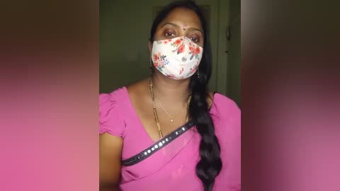 Media: Video of a South Asian woman with medium skin tone, dark hair in a single braid, wearing a pink top, floral mask, and black crossbody bag, in a dimly lit indoor setting.