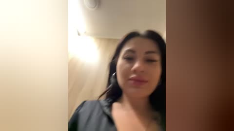 Media: A blurry video of a Latina woman with long dark hair, wearing a black blouse, standing in a dimly lit hallway with beige walls and a light fixture.