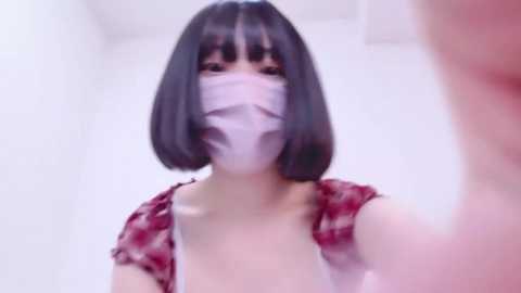 Media: A blurry video of an East Asian woman with straight black hair, wearing a red plaid shirt and a white face mask, standing indoors against a white background.