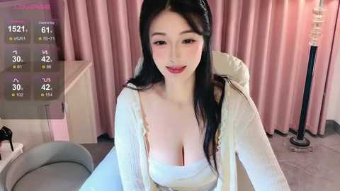 Media: A video of a young Asian woman with long black hair, fair skin, and a curvaceous figure, wearing a low-cut white dress, sitting in a modern room with pink curtains and a digital clock.