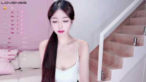 Media: Video of an East Asian woman with long black hair, wearing a white tank top, sitting on a beige couch in a modern, light-pink room.