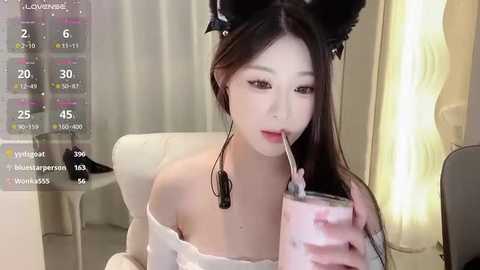 Media: Video of an East Asian woman with fair skin and long dark hair, wearing black cat ears and a white off-shoulder top. She's drinking a pink milkshake with a straw, in a modern, brightly lit room.