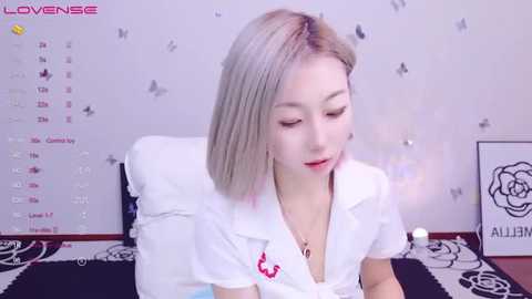 Media: A video of a young Asian woman with light skin, straight blonde hair, and red lipstick, wearing a white shirt with a red bow, seated in front of a calendar with butterfly decals and a framed flower.