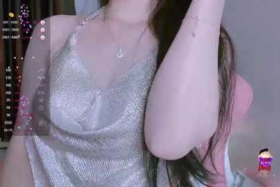 Media: Video of a light-skinned woman with long brown hair, wearing a silver sequined halter top, sitting with one arm raised. Background is blurred with social media icons and text.