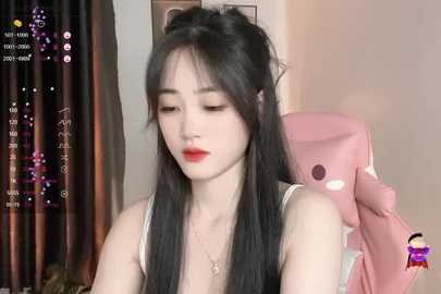 Media: Video of a young Asian woman with long black hair, wearing a sleeveless top, sitting indoors. She has fair skin and red lipstick. Background includes a pink plush toy and a blurred window.