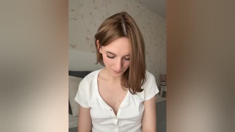 Media: Video of a young Caucasian woman with light skin and shoulder-length brown hair, wearing a white, button-up shirt, sitting in a bedroom with white floral wallpaper and a bed in the background.