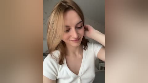 Media: Video of a young Caucasian woman with light skin and shoulder-length, light brown hair, wearing a white, short-sleeved cardigan. She has a soft smile and is gently touching her hair, with a blurred background.