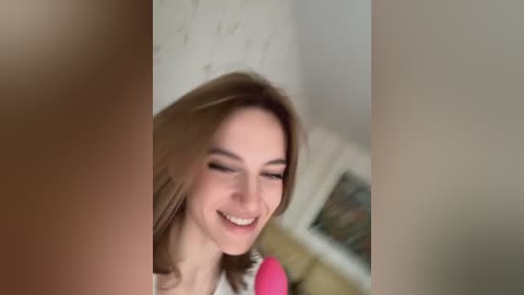 Media: Video of a young Caucasian woman with light skin and shoulder-length brown hair, smiling with eyes closed, wearing a pink top. Background features a blurred, white-walled room with a beige baseboard and a window.